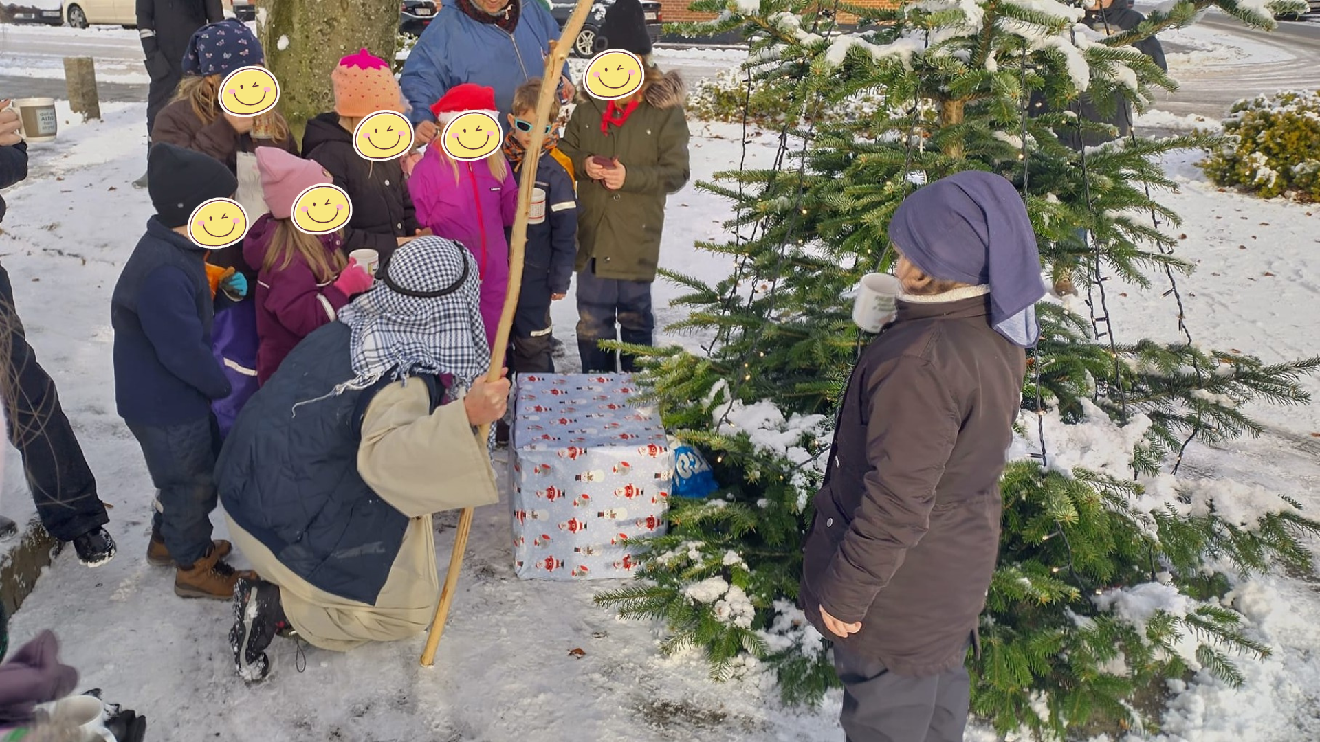 Hyrde sidder på jorden med en stav foran et juletræ. Børneflok står rundt omkring.